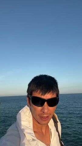 A man wearing sunglasses and a white hoodie stands facing the camera with the ocean and clear blue sky in the background. The man is looking directly at the camera. There is a sailboat visible in the distance on the left side of the frame.