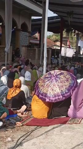 A group of people, many of whom are women wearing hijabs and long dresses, are gathered outdoors. They are sitting on the ground on rugs. In the foreground, a woman is using a purple umbrella to shield herself from the sun. In the background, there appears to be a building with an arched entryway, possibly a mosque, and tents set up, suggesting an outdoor event or gathering. Some individuals are standing near the building. The ground is a mixture of dirt and concrete. The lighting suggests it is daytime, possibly a sunny day.