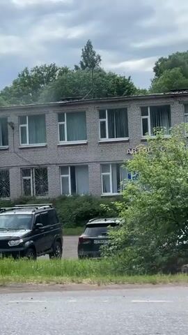 A grey brick building stands behind a road and a patch of grass. The building has multiple windows and a sign on the front that reads "ШКОЛА ДОСААФ". A black SUV is parked to the left of the building. A blue minivan is parked to the right of the building. There are trees behind the building and a cell tower on top of the building. The sky is cloudy.
