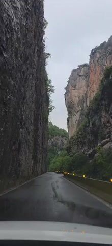 The video shows a car driving on a wet road through a narrow canyon. The canyon walls are tall and made of gray and reddish-brown rock. There is green vegetation growing on the canyon walls. The sky is overcast. The car is moving forward at a moderate speed. There are yellow reflective barriers lining the side of the road.