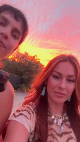 The video shows three young women, likely friends, taking a selfie video outdoors at dusk. The woman closest to the camera is in the foreground, and has long red hair, and a tan and black animal print top. Her friends are to her left and just behind her. The woman on the left has short dark hair, and is wearing a black top. The woman behind the main subject, and to her right, has long light brown hair, and is wearing a green top. They are all smiling and appear to be in good spirits. The woman behind and to the left waves her arm with a wristband on it. The background is blurred, but appears to be a road with cars and trees. The sky is a gradient of orange and pink, indicating it is either sunrise or sunset.