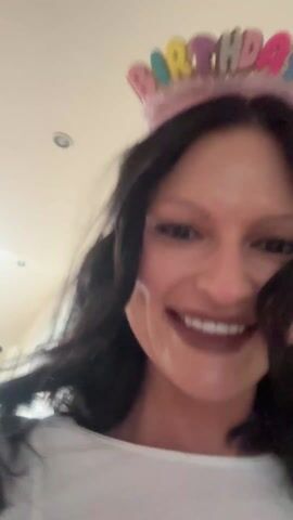 A woman wearing a white top and a birthday headband smiles at the camera. A man plays the saxophone in the background. The woman turns towards the man and moves her hand slightly towards him. The woman then turns back to the camera and continues to smile. The camera is moving slightly throughout the video. There is a picture frame on the wall. The background is a room with white walls.