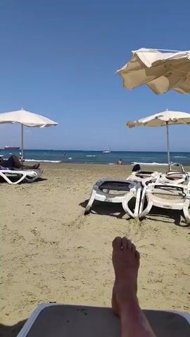 The video shows a beach scene on a sunny day. In the foreground, a person's bare feet are visible, resting on a lounge chair. Two white beach umbrellas are partially visible at the top of the frame. In the middle ground, there are several empty lounge chairs on the sandy beach. The sea is in the background, with gentle waves rolling onto the shore. A sailboat is visible in the water, and a person is sitting on the sand near the waterline. Another person is swimming in the sea. Further in the distance, a large ship is anchored offshore. The sky is clear and blue.