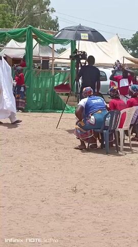 The video shows a scene that appears to be an outdoor event with several people. A man in a white robe walks slowly in the foreground with a cane.  To the right, a group of women are seated in chairs, some wearing head coverings and red or patterned tops. A cameraman with an umbrella stands behind them, filming. In the background, there are tents, greenery, and vehicles visible. The ground is dusty.  Some people are standing and watching the event unfold. The overall impression is of a ceremony or celebration taking place.