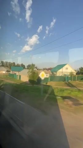 This is a video taken from a moving vehicle, likely a bus or train, looking out a window. The view captures a rural or suburban landscape under a clear blue sky with scattered white clouds. The foreground shows a road and green grass, with reflections on the window glass. In the midground, there are several houses with green roofs, light-colored walls, and fenced yards. Some trees and utility poles are also visible. The scene appears to be a sunny day, and the motion of the vehicle creates a slight blur effect.