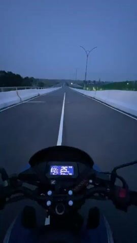 The video captures a first-person perspective from a motorcycle rider traveling on a straight, multi-lane highway during dusk or early night. The motorcycle's illuminated dashboard is prominently visible, displaying the speed, which ranges from 27 km/h to 41 km/h, along with the gear indicator (showing 'N', then '1', '2', '3'), a fuel gauge, and an odometer reading (partially visible, around 3130 km). A "Honda" logo is visible on the dashboard.

The asphalt road features a clear white center line and white shoulder markings, flanked by concrete barriers on both sides. Tall, distinctive Y-shaped streetlights are spaced along the road, their lights visible in the distance. The sky is a dark blue, indicating the transition from twilight to night. Trees and vegetation line the horizon, suggesting an open landscape. The ride appears smooth and uninterrupted on the clear road. This scene takes place in Rupganj, Bangladesh.