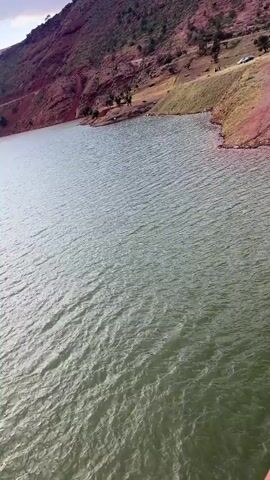 The video shows a view of a lake from a bridge. The water is green and has small waves. The shoreline is visible on the right and left of the frame, with red soil and sparse vegetation. A hill rises in the background. The sun shines through the clouds, creating a reflection on the water. The sky is partly cloudy. A white and red railing is seen in the bottom left, indicating the view is from a bridge.