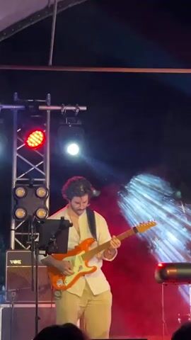 A man with dark hair is playing an orange electric guitar on a stage. He is wearing a white button-down shirt and light-colored pants. There are stage lights and equipment in the background, including a speaker and a light rig with red and white lights. The scene appears to be an outdoor performance at night.