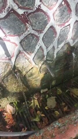 The video opens with a close-up of a stone wall with white grout. Water is seen dripping from spouts embedded in the wall onto a grate covered with fallen leaves. The camera then pans to the right, revealing more of the stone wall and a dark hose coiled to the left. A wooden chair sits in front of the wall, and further to the right, a tree grows against the stone structure. The scene shifts to a stone pathway leading upwards, flanked by a stone wall on the left and vegetation on the right. Sunlight filters through the leaves, casting shadows on the path. The video concludes with a view of the ascending stone steps.