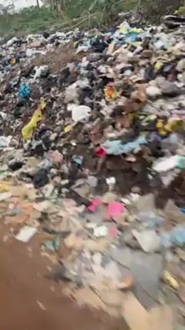 The video depicts an expansive area heavily littered with trash. The debris covers the ground, stretching up a small hill. Amidst the garbage, there are hints of fabric, plastic bags, and various unidentifiable objects. The trash includes dark and light colors, suggesting a mix of materials. In the background, there are trees including palm trees, along with other greenery, hinting at a tropical environment. The sky appears overcast, suggesting a cloudy day. Overall, the scene portrays a severe environmental issue with widespread waste accumulation.