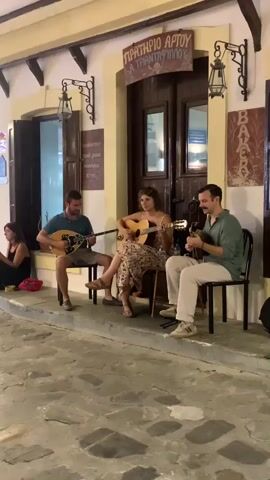 Three musicians, two men and one woman, are seated on chairs outdoors in front of a building that appears to be a bakery. The woman in the center is playing an acoustic guitar and singing. To her left, a man plays a bouzouki, and to her right, another man plays a mandolin. A woman is seated to the far left of the frame, out of focus. The ground is paved with irregular stones. The building has a traditional facade with wooden doors and windows, and signs in Greek script are visible above the entrance. The lighting suggests it is either evening or early morning.