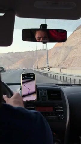 The video shows the interior of a car from the perspective of a passenger. The driver's left hand is on the steering wheel. A smartphone mounted on the dashboard displays a navigation app with a route. The driver's face is visible in the rearview mirror, looking forward. The car is driving on a highway with mountainous, arid terrain on the right side. The sun appears to be setting, casting a warm light. There is a metal guardrail along the highway, and power poles are visible in the distance. The car radio is visible below the phone. The video is in landscape orientation.