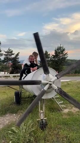 A man and a woman are sitting on the wing of a small, grounded airplane. The man is in the front, holding onto a control lever that is connected to the propeller. He is smiling and looking at the camera. The woman is behind him, looking down. The airplane is white with a large, four-bladed propeller. The propeller is tilted, suggesting it might be a prop or is in motion. The scene is set outdoors, with green grass and trees in the background. The sky is partly cloudy with a sunset visible. The overall impression is that of a playful or staged scene.