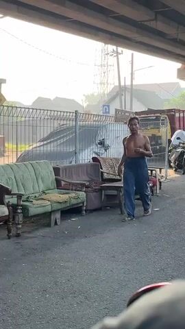 The video shows an outdoor scene under an overpass. A shirtless man wearing blue pants walks across an asphalt surface. To his left, several pieces of furniture, including two couches and a table, are placed on the ground. Behind the furniture, a metal fence runs horizontally. In the background, there are vehicles, including a truck and a white vehicle that appears to be a motorcycle, as well as buildings and utility poles. The lighting suggests it is daytime, possibly morning or late afternoon, with soft sunlight filtering through.