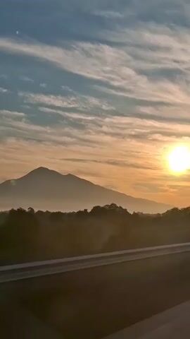 This video shows a sunrise over a mountainous landscape. The sun is bright and partially obscured by clouds, casting an orange glow over the sky. A mountain range is visible in the distance, silhouetted against the bright sky. In the foreground, there is a road or highway, with a guardrail separating the viewer from the scenery. Trees and other vegetation line the roadside, shrouded in a light mist. The video appears to be filmed from a moving vehicle, likely a car or bus, as indicated by the slight motion blur and the perspective.