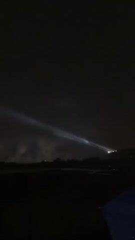 The video is a nighttime scene. In the sky, there is a bright spotlight beam moving. Then, fireworks begin to explode in the dark sky, creating bright bursts of light. The fireworks appear to be occurring over a treeline, with the lower part of the frame filled with darkness, suggesting the viewer is at ground level.