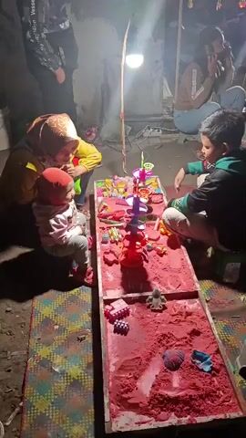 A group of people are gathered around a table in a dimly lit, indoor space. A young child in a red hat and pink sweater is sitting and looking at toys on the table. To their left, an adult wearing a yellow jacket and a hijab is also looking at the toys. Across from them, a boy in a green and black jacket is playing with the toys. The table is covered with a variety of colorful toys and red sand, with sandcastle molds and small toy figures visible. The environment appears to be a makeshift play area, possibly outdoors or in a poorly lit room, with a bare bulb hanging overhead providing illumination. Other individuals are present in the background, partially obscured by shadows.