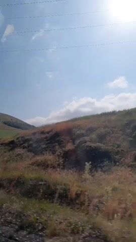A video of a landscape, likely taken from a moving vehicle. The scene consists of rolling hills covered in dry grass and some sparse vegetation. The sky is blue with scattered white clouds, and the sun is visible, creating a bright glare in some parts of the footage. There are several tall, slender structures that appear to be telecommunication towers or power line pylons situated on the crest of the hills. The foreground is blurred, suggesting motion. There are no people or animals visible in the video.