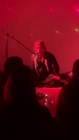 Here is a concise description of the video:

A person, presumably a singer or musician, is sitting cross-legged on a stage illuminated by red lighting. They are singing into a microphone, which is positioned in front of them. The performer is wearing dark clothing. In the foreground, the backs of heads of audience members are visible. The lighting creates a hazy and dramatic atmosphere.