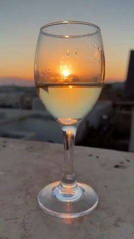A stemmed wine glass, partially filled with a pale yellow liquid, stands on a textured surface. The glass is positioned in the foreground, with its base resting on what appears to be a stone ledge or balcony railing. The liquid inside the glass is illuminated by the setting sun, which is visible through the glass, creating a warm, golden glow. The sun itself is a bright disc, appearing just above the horizon line, and its light is refracted and reflected by the liquid and the droplets of condensation on the outside of the glass.

The background shows a blurred landscape of buildings and rooftops, suggesting an urban or suburban setting. The sky transitions from a soft orange and yellow near the horizon, indicative of sunset, to a dusky blue higher up. The overall mood is serene and peaceful, amplified by the warm light and the tranquil scene. The time of day is clearly sunset. There is no visible text in the media file. The location is indicated as Katzrin, Israel.