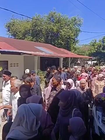A crowd of people, predominantly women wearing hijabs, are gathered outdoors in front of a building with a red corrugated metal roof. The sky is clear and blue, and trees are visible in the background. Many people are looking in the same direction, and some appear to be walking. The scene suggests a communal gathering or event.