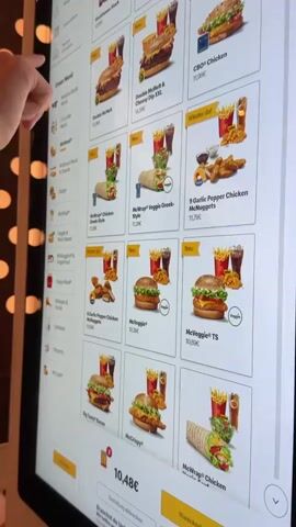 The video shows a close-up of a digital menu screen at a McDonald's. The screen displays various food items with images and prices. The menu is in German, with categories like "Unser Menü" (Our Menu) and "Wieder da!" (Back again!). Several food items are visible, including burgers, wraps, chicken nuggets, and fries. Some items are marked as "Neu" (New). The prices are displayed in Euros. The screen appears to be a touchscreen interface. The background is blurred, but suggests a dimly lit restaurant environment. There are no people clearly visible in the video. The overall impression is a view of a modern fast-food ordering system.