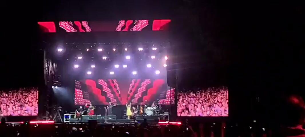A wide shot shows a live outdoor concert. A band performs on a brightly lit stage with large screens displaying images of the performers. A large crowd of people is visible in the foreground, facing the stage. The overall lighting is dim, with stage lights dominating the scene. The video is grainy and low-resolution.