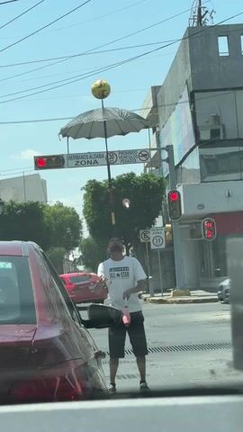 The video shows a street performer juggling at a traffic stop in an urban environment. The performer, wearing a white "Converse All-Star" t-shirt and dark shorts, juggles pink juggling clubs while balancing a soccer ball on top of an open umbrella mounted on a pole. The scene takes place at a traffic intersection with multiple cars waiting at a red light. Traffic signs are visible in the background, indicating speed limits and street names. A red car is partially visible in the foreground, presumably the location from where the video is recorded. The weather appears to be sunny and clear.