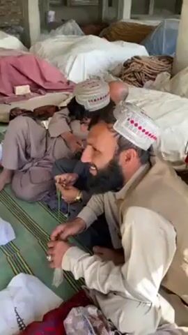 Several men sit on a green striped surface in a cluttered indoor space with white bags or bundles stacked behind them. The man closest to the camera has a full black beard, a white cap with a red pattern, and wears a tan outfit. He looks directly at the camera and smiles. He holds a strand of beads in his hands. Another man to his left has a similar cap and beard. The environment is filled with piled materials, suggesting a warehouse or storage area.