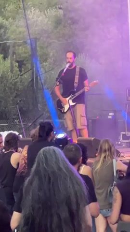 This media file captures a live musical performance at an outdoor event, likely a concert or festival, in Analiontas, Cyprus. The scene is set at dusk or night, with purple and blue stage lights illuminating the performers and a portion of the audience. The backdrop suggests an open area with trees visible in the upper left, hinting at a rural or semi-rural location.

The primary subject is a male guitarist and singer on stage, dressed in a black t-shirt and khaki shorts. He is actively playing his electric guitar and singing into a microphone. To his right, on stage, are Marshall amplifiers, typical for live music setups. Another guitarist, a heavier-set male in a white t-shirt and black shorts, is visible further back on the right side of the stage.

In the foreground and mid-ground, a diverse audience is gathered, facing the stage. Their backs are mostly to the camera, but their presence indicates an engaged crowd. Some individuals have their hair tied up, and others have long, flowing hair. The lighting creates a vibrant, energetic atmosphere. The overall mood appears to be one of enjoyment and immersion in the live music experience. The visible text on the amplifiers reads "Marshall".