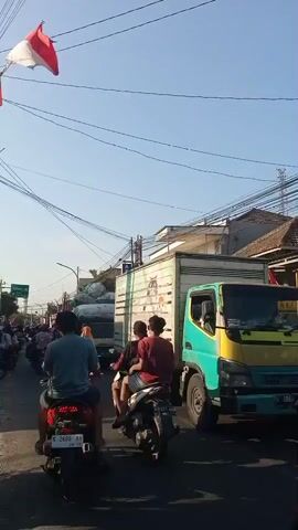 A traffic jam is depicted on a sunny day, with several motorbikes and a truck visible. An Indonesian flag, red and white, is flying in the upper left corner. The road appears to be paved, and there are buildings and power lines along the sides. Several people are riding on the motorbikes, some are wearing helmets, and one person is wearing a headscarf. The truck is green and white, and it appears to be carrying goods. The vehicles are moving slowly due to the traffic.