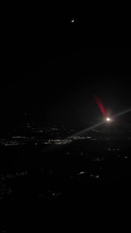 The video shows a nighttime view from an airplane window. Below, city lights twinkle in the darkness. The plane's wing, illuminated in red by the aircraft's lights, is visible. A crescent moon shines in the upper portion of the frame.