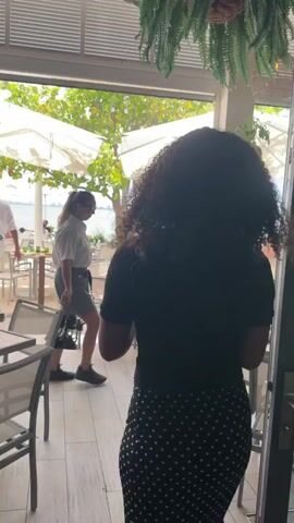 A woman with curly dark hair, wearing a black top and a polka dot skirt, stands with her back to the camera. She is holding a menu. She is in an outdoor dining area of a restaurant. In the background, there are tables and chairs, large white umbrellas providing shade, and a view of the water with a city skyline in the distance. Other diners are visible in the background. Lush green plants are placed throughout the dining area.