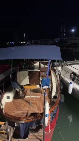 The video shows a nighttime view of a marina with several boats docked. The camera pans from left to right, focusing on a wooden-decked boat in the foreground, then moving to a white cabin cruiser next to it, and finally showing a wider view of other boats in the background. The water is calm and reflects the lights from the boats and the distant shore. The sky is dark, indicating it is night, and a faint moon can be seen in the upper right portion of the frame. The boats appear to be moored and unattended. The overall scene is peaceful and serene, highlighting the quiet atmosphere of a harbor at night.