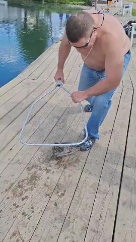 A man kneels on a weathered wooden pier beside a body of water in Kipen, Russia. He is shirtless, wearing blue jeans and sunglasses, and appears to be focused on a fish he has caught. The man holds a large, metal-framed landing net, which contains a sizable trout.

The fish, dark with distinctive pinkish-orange spots along its side, lies within the netting on the wooden planks of the pier. The man's right hand is gently holding the fish near its mouth, where a fishing hook or lure is visible. He seems to be in the process of unhooking his catch.

The scene is set outdoors during daylight hours, indicated by the bright light. The water in the background is calm, reflecting the surroundings. The overall atmosphere suggests a casual fishing outing. There is no discernible text visible in the video.