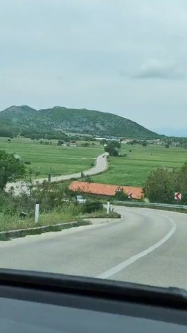 A car is driving along a winding road through a rural landscape. The road has a white line down the center and guardrails on the side. There are red and white signs indicating curves in the road. In the distance, there is a green field with scattered trees and a low mountain range in the background. The sky is overcast. A silver car passes in the opposite direction. The view is from the driver's perspective, looking out through the windshield.