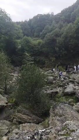 The video shows a rocky, wooded ravine with a waterfall cascading down a mossy rock face. Several people are visible in the distance, navigating the rocky terrain. One person, a man wearing a dark t-shirt with a yellow stripe and grey pants, is standing on a large rock in the foreground, facing away from the camera and towards the waterfall. The weather appears overcast.