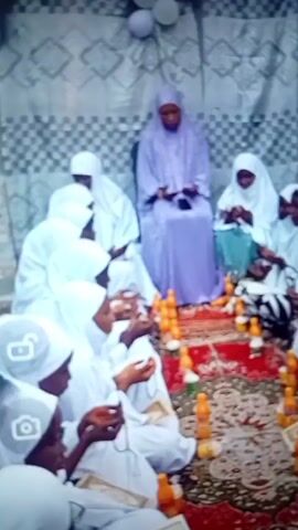 Several people wearing white hijabs are gathered on a carpet, some holding objects in their hands. One person is wearing a light purple robe and sits behind them. There are orange bottles on the carpet. The background features a patterned fabric. Some individuals appear to be praying or engaged in a religious ceremony.