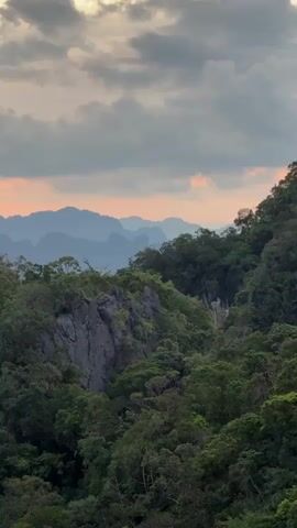 A static medium shot captures a mountain landscape at dusk. The foreground is dominated by dense green foliage, with a prominent gray rock formation on the left. In the midground, rolling hills covered in trees stretch into the distance. The background features layers of hazy, blue-toned mountains silhouetted against a sky filled with dramatic clouds. The setting sun casts an orange glow through the clouds, creating a warm light on the horizon. The overall impression is one of a serene, natural environment bathed in the soft light of twilight.