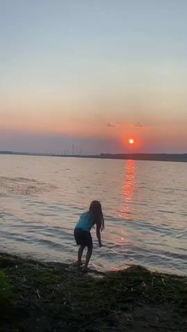 A child kneels at the edge of a body of water as the sun sets. The sun is a bright orange orb low on the horizon, casting a shimmering reflection on the water. The sky is a gradient of soft pinks and blues. The child is wearing a blue shirt and dark shorts. They are reaching a hand into the water. The shore is covered in dark dirt and green moss. The scene appears peaceful.