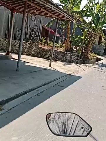 The video shows a point-of-view shot from a motorcycle traveling down a paved road in what appears to be a village or rural area. On the left side of the road, there are houses with banana trees and other vegetation. There are also small shops and businesses visible, including a shop selling lumber and a store with drinks displayed outside. The road is bordered by a concrete drain. The weather appears sunny and clear. The video is very short and appears to be a segment from a longer recording.