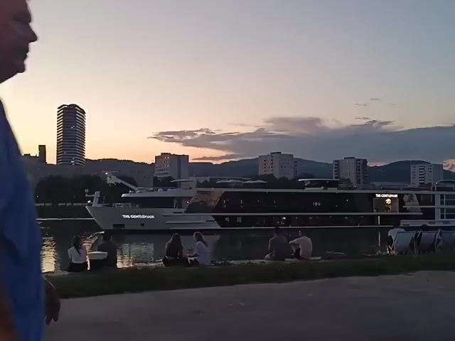 The video begins with a close-up of two people walking away from the camera. The person on the left is wearing a pink shirt, and the person on the right is wearing a blue t-shirt. As the video progresses, the camera pans to the right, revealing a wider view of a riverfront scene at dusk. A large white cruise ship is docked on the river. Several people are sitting on the grassy bank of the river, some appearing to be couples. In the background, there is a cityscape with several buildings, including a tall, slender skyscraper. The sky is a soft gradient of orange and purple, indicating either sunrise or sunset. The overall atmosphere is calm and peaceful.