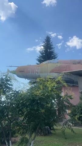 A static, medium shot, filmed outdoors under a bright blue sky with scattered clouds, shows an old, weathered fighter jet parked on a concrete pedestal. The jet is painted in a camouflage pattern of faded reds, greens, and yellows, with the number 509 visible on its nose. Green trees and bushes partially obscure the aircraft. In the background, a house with a pink wall and a white car are visible. In the foreground, pink roses bloom in the grass. The video pans slightly from left to right, revealing more of the jet's wing and landing gear. There are no people visible in the video.