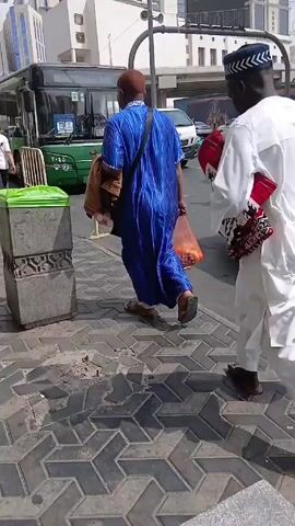 A bright outdoor shot depicts several individuals walking along a brick-paved sidewalk adjacent to a green and white bus. The bus is parked along the curb with its doors closed. Two prominent figures, a man in a blue robe and a man in a white outfit with a black and white cap, are walking in the same direction, away from the camera. The man in blue carries a brown bag and a plastic bag of oranges, while the man in white has a patterned red bag and a black shoulder bag. The sidewalk is lined with metal barricades and green-lidded trash cans. In the background, tall buildings with reflective windows can be seen, including signs indicating "Marriott" and "Hilton". Other people in traditional attire are walking in the same direction, and cars are visible on the street parallel to the sidewalk. The overall scene suggests a bustling urban environment with people going about their day.