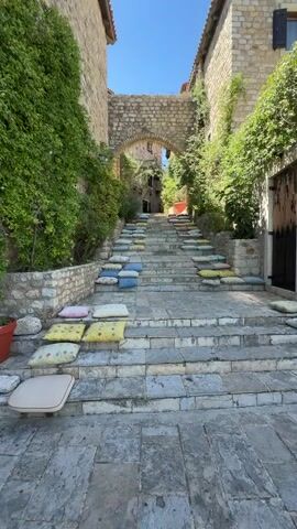 The video shows a stone staircase leading upwards between old stone buildings. The steps are adorned with numerous colorful cushions, arranged on each step. Lush green foliage grows along the stone walls on both sides of the staircase, and an arched passageway is visible at the top. The scene is bathed in bright sunlight, suggesting it is daytime. There are no people visible in the video.