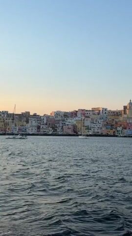 The video shows a view of the island of Procida from the sea, with the colorful houses and buildings of the town stretching along the coastline. The water in the foreground is dark and slightly choppy, and there are two sailboats anchored in the bay. The sky is clear and blue, with a slight golden hue near the horizon.