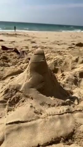 Here's a description of the video:

The video shows a close-up of a small sandcastle on a beach. The sandcastle is simple, shaped like a cone, and placed on a bed of dry sand. In the background, the ocean is visible with gentle waves. A person is seen standing in the water in the distance. The sky is blue and clear.