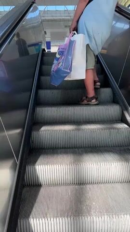 The video shows a person standing on an escalator. The person is wearing a light blue dress and olive green shorts. They are holding two shopping bags, one blue with a picture on it and one white. The escalator is made of metal with ribbed steps. The background appears to be a large indoor space, possibly a train station or mall, with bright lighting and some signage visible. The person is facing away from the camera and appears to be moving upwards on the escalator.