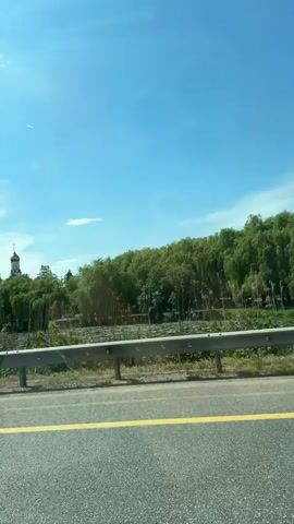 The video captures a scenic view from a moving vehicle on a clear, sunny day. The camera looks out over a road with yellow dividing lines and a metal guardrail. Beyond the guardrail, a landscape of lush green trees, possibly willows, stretches across the midground. In the distance, two distinct Orthodox Christian structures are visible. One is a tall, white bell tower with a prominent golden dome and cross. Further to the right, a second building with a blue dome and cross can be seen, partially obscured by the trees. The sky above is bright blue with a few scattered clouds. Towards the end of the clip, the rear of a light-colored sedan briefly passes into view from the left, indicating other traffic on the road. The accompanying audio includes speech in Russian.