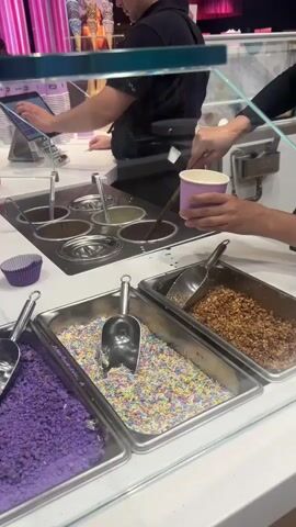 This video shows the interior of an ice cream shop. A person is holding a purple cup and a scoop that is dipping into a vat of what appears to be melted chocolate. Another person, wearing a black shirt and apron, is standing behind a counter, possibly preparing an order or operating a tablet. In front of the counter are three large metal trays filled with toppings: the leftmost tray contains purple sprinkles, the middle tray has rainbow sprinkles, and the rightmost tray holds a brown, crumbly topping. Behind the counter, there are more ice cream cones and a sign that says "VICTORIA'S". The lighting is bright, and the overall environment appears clean and modern.